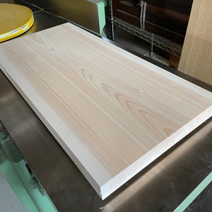 May include: A large, light-colored wooden board with a smooth surface and a slightly rounded edge. The board is resting on a metal surface.