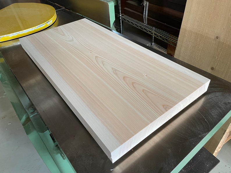 May include: A large, light-colored wooden board with a smooth surface and a slightly rounded edge. The board is resting on a metal surface.