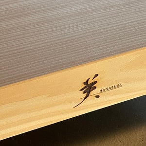 May include: A light brown wooden cutting board with a gray top surface. The board has a logo engraved on the side that says "HANABUSA" with a Japanese character.