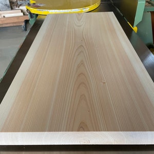 May include: A large, light-colored wood plank with a smooth surface and visible grain patterns. The plank is lying flat on a dark surface.