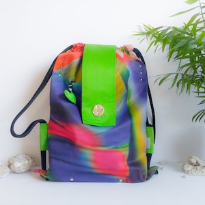 Può includere: Zaino con coulisse con motivo astratto nei toni del viola, rosa e verde. La borsa presenta una patta verde brillante fissata con un bottone di legno. Cinghie in corda nere e dettagli verdi. L'altezza è di circa 40 cm.