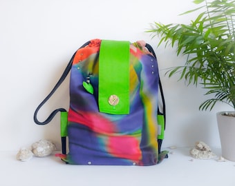 Può includere: Zaino con coulisse con motivo astratto nei toni del viola, rosa e verde. La borsa presenta una patta verde brillante fissata con un bottone di legno. Cinghie in corda nere e dettagli verdi. L'altezza è di circa 40 cm.