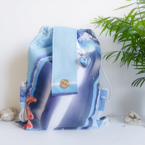 Könnte beinhalten: Ein blau-weiß gemusterter Rucksack mit einem passenden hellblauen Handtuch. Der Rucksack hat einen Kordelzugverschluss und einen kleinen herzförmigen Anhänger. Auf dem Handtuch befindet sich ein Holzknopf. Der Rucksack ist etwa 40 cm hoch.