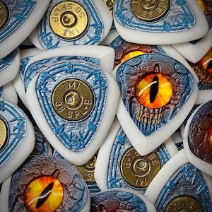 May include: A collection of triangular guitar picks featuring two distinct designs. One design displays a brass 38 SPL bullet casing centre, while the other features a realistic yellow and orange reptilian eye set within a textured blue frame.