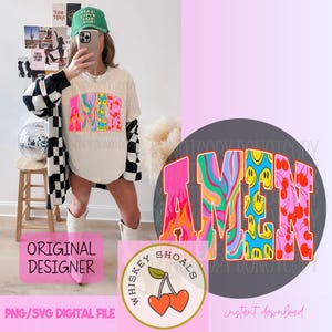 Puede incluir: Camiseta color crema con la palabra "AMEN" en un dise&ntilde;o retro colorido. Las letras presentan una mezcla de patrones, incluyendo llamas, caras sonrientes y cerezas. Tambi&eacute;n se ve una gorra verde y una chaqueta a cuadros en blanco y negro.