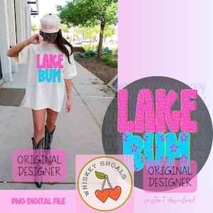 Puede incluir: Camiseta color crema con las palabras "LAKE BUM" en rosa y azul, con estrellas y lunares. La imagen incluye un logotipo de "Whiskey Shoals" con cerezas y el texto "ORIGINAL DESIGNER".