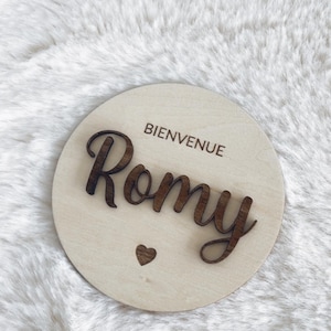 May include: A round wooden sign with the text "Bienvenue Romy" in brown lettering. A small heart is below the name.