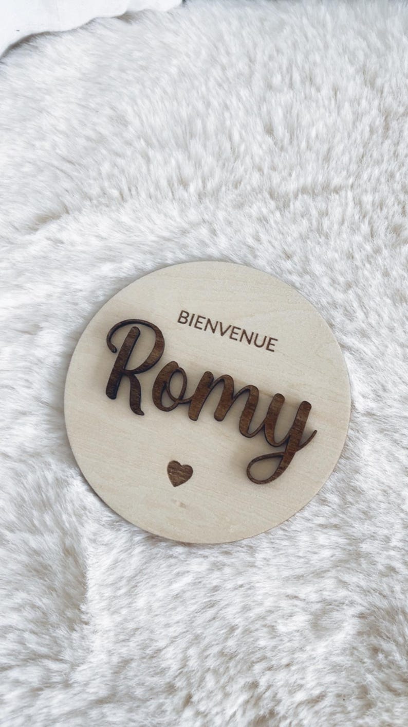 May include: A round wooden sign with the text "Bienvenue Romy" in brown lettering. A small heart is below the name.