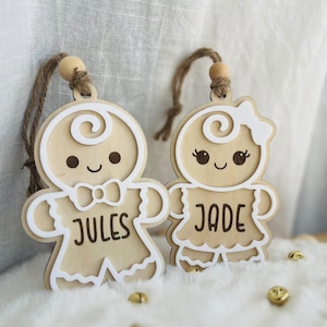 Peut inclure: Deux décorations en bois en forme de bonhomme en pain d'épice avec des contours blancs et des attaches en ficelle de jute. L'une porte un nœud papillon et le nom JULES, l'autre un nœud dans les cheveux, un col à volants et le nom JADE.
