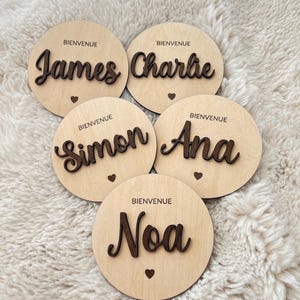 May include: Five wooden round plaques with the words "Bienvenue" and names "James", "Charlie", "Simon", "Ana", and "Noa" written in cursive.