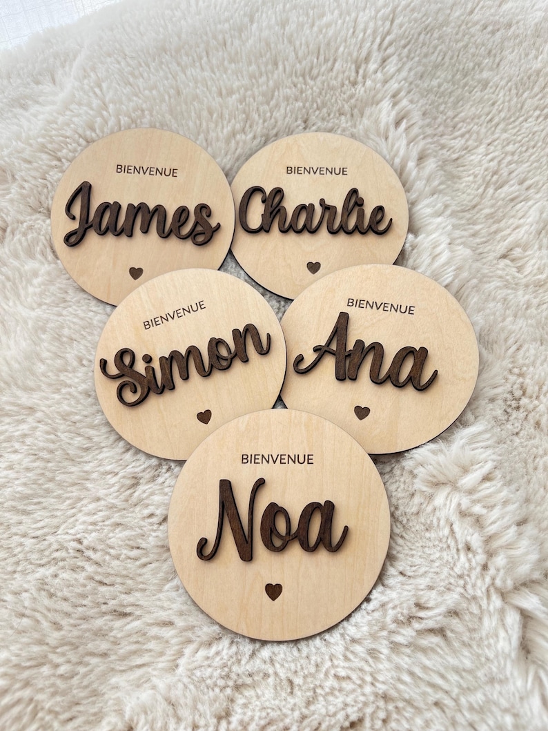 May include: Five wooden round plaques with the words "Bienvenue" and names "James", "Charlie", "Simon", "Ana", and "Noa" written in cursive.