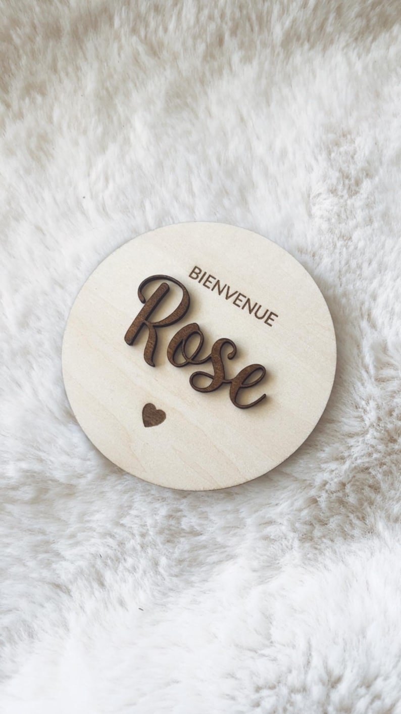 May include: A round wooden sign with the text "Bienvenue Rose" in brown lettering. A small heart is carved into the wood below the text.