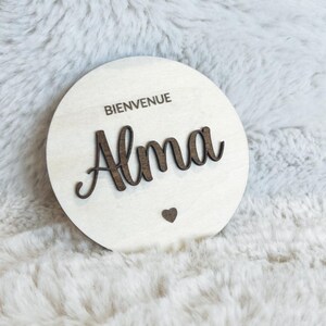 May include: A round wooden sign with the text "Bienvenue Alma" in brown lettering. A small heart is below the name.