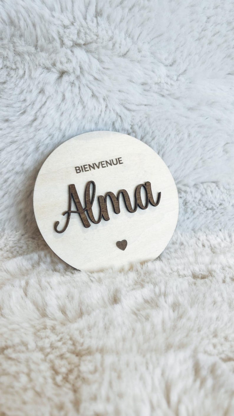 May include: A round wooden sign with the text "Bienvenue Alma" in brown lettering. A small heart is below the name.