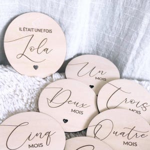 Peut inclure: Un ensemble de disques de naissance circulaires en bois clair avec des inscriptions gravées en noir. Les disques portent les mentions "Il était une fois Zola", "Un mois", "Deux mois", "Trois mois", "Quatre mois" et "Cinq mois" avec des petits cœurs.