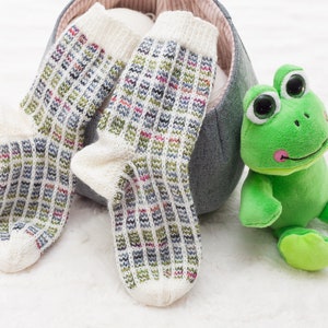 Könnte beinhalten: Zwei Paar weiße Stricksocken mit einem bunten geometrischen Muster. Die Socken befinden sich in einem grauen Korb mit einem grünen Plüsch-Frosch.