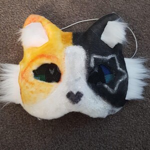 May include: A handmade cat mask with a split design. One side is orange and yellow, the other black. Both sides have mesh eye coverings and white fur accents. A small black heart is on the nose.