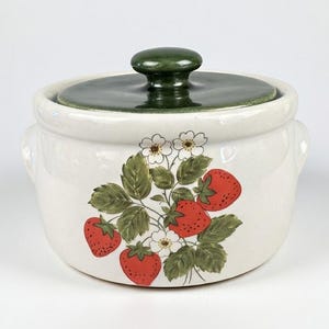 May include: A white ceramic lidded jar with a dark green knob. The jar is decorated with a strawberry and leaf design. The lid is dark green. The jar has two small handles on either side.