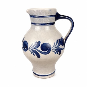 May include: A light gray ceramic pitcher with a bulbous body and a single handle. The pitcher features dark blue hand-painted floral scroll patterns around the center, with horizontal blue bands encircling the neck and the base of the vessel.