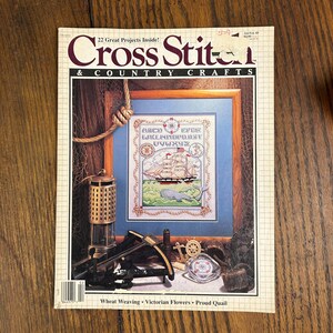 May include: A vintage issue of "Cross Stitch & Country Crafts" magazine. The cover features a framed cross-stitch design of a sailing ship, whale, and alphabet. Additional craft-related items are arranged around the frame.