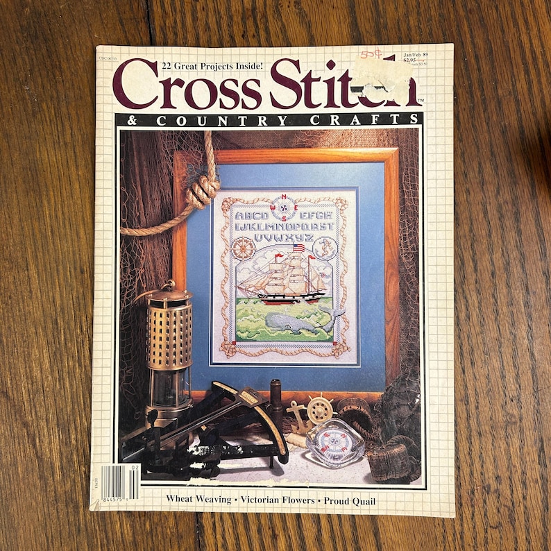 May include: A vintage issue of "Cross Stitch & Country Crafts" magazine. The cover features a framed cross-stitch design of a sailing ship, whale, and alphabet. Additional craft-related items are arranged around the frame.