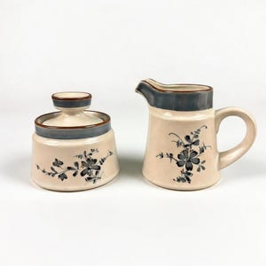 May include: A cream-colored ceramic creamer and sugar bowl set. The set features a floral design in dark gray and blue. The rims and lids are accented with a dark blue trim. The sugar bowl has a lid with a knob.