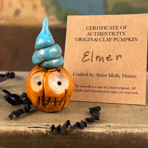 May include: A small orange clay pumpkin figurine with a blue spiral stem and carved facial features, accompanied by a tan certificate of authenticity card that reads: CERTIFICATE OF AUTHENTICITY ORIGINAL CLAY PUMPKIN Elmer Crafted by Artist Molly Mattin.