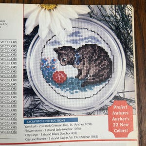 May include: A cross-stitch pattern featuring a brown tabby cat with a blue collar, gazing at a red yarn ball. The design includes blue flowers and a blue sky, all within a circular frame. The text reads "Project features Anchor's 22 New Colors!"