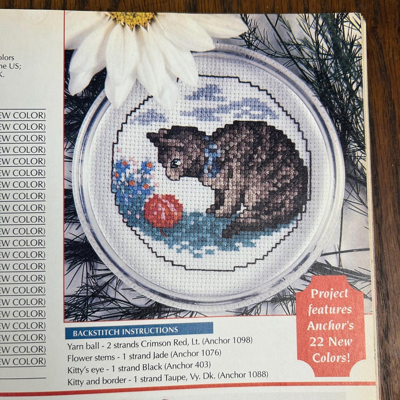 May include: A cross-stitch pattern featuring a brown tabby cat with a blue collar, gazing at a red yarn ball. The design includes blue flowers and a blue sky, all within a circular frame. The text reads "Project features Anchor's 22 New Colors!"