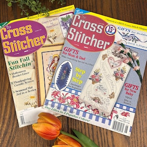 May include: A collection of vintage "Cross Stitcher" magazines. The covers feature colorful cross-stitch patterns, floral designs, and text such as "Fun Fall Stitchin'" and "Gifts for Mom & Dad." A tulip rests in the foreground.