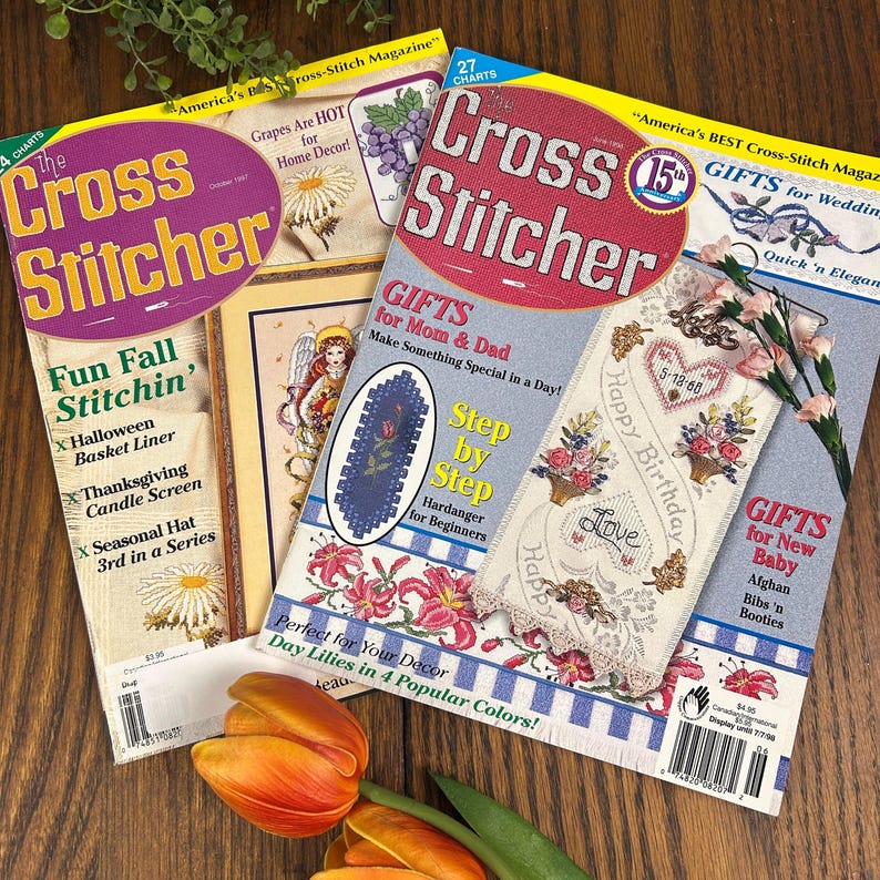 May include: A collection of vintage "Cross Stitcher" magazines. The covers feature colorful cross-stitch patterns, floral designs, and text such as "Fun Fall Stitchin'" and "Gifts for Mom & Dad." A tulip rests in the foreground.