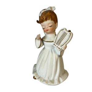 May include: A vintage porcelain figurine of a girl in a white dress, holding a harp. The figurine has brown hair, closed eyes, and a bow. Gold trim accents the dress and harp. The figurine is a decorative item.