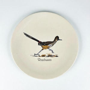 May include: A cream-colored ceramic plate featuring a detailed illustration of a roadrunner bird in motion. The bird is depicted with brown, black, and tan feathers, a long tail, and a distinctive crest. The word "Roadrunner" is printed below the bird.