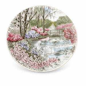 May include: A round ceramic plate featuring a detailed illustration of a garden scene with a pond, a small wooden bridge, and lush clusters of pink, purple, and white flowers. The artwork uses fine black lines and soft watercolor-style shading.