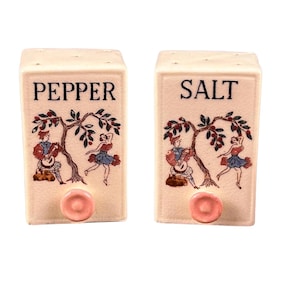 May include: A pair of ceramic salt and pepper shakers. The rectangular shakers are off-white with the words "PEPPER" and "SALT" in black. Each shaker features a tree with figures playing instruments and dancing. Pink knobs are at the bottom.
