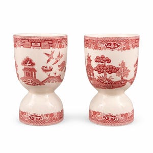 May include: Two white ceramic egg cups with a red Chinoiserie design. The design includes pagodas, bridges, birds, and trees. The egg cups have a unique hourglass shape and are suitable for serving eggs.