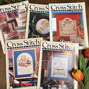 May include: A collection of vintage "Cross Stitch & Country Crafts" magazines. The covers feature cross-stitch designs, including framed samplers, teddy bears, and a ship. The magazines are stacked on a wooden surface, with a few flowers in the corner.