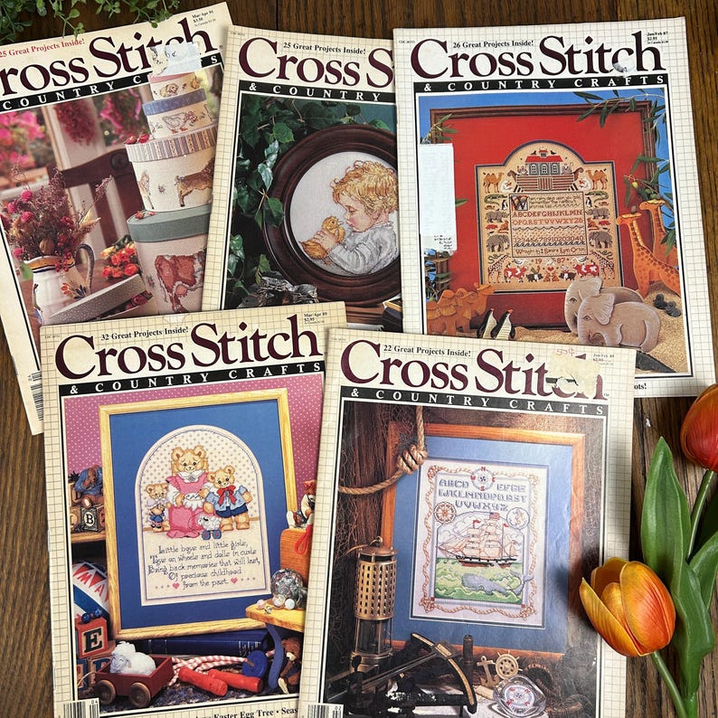 May include: A collection of vintage "Cross Stitch & Country Crafts" magazines. The covers feature cross-stitch designs, including framed samplers, teddy bears, and a ship. The magazines are stacked on a wooden surface, with a few flowers in the corner.