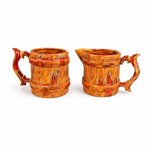 May include: Two ceramic mugs with a wood-grain design. The mugs are brown with red accents and ornate handles. One mug has a spout. The mugs are likely for serving beverages.