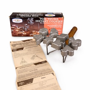 May include: Vintage Tom Thumb Instant Cookies set. The set includes a box, instructions, and a metal cookie mold with various shapes. The box features the product name and images of the cookies. The mold has a wooden handle and is designed for making bite-sized cookies.