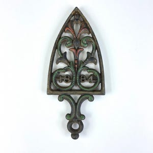 May include: An antique cast iron trivet with an ornate design. The trivet is shaped like an iron and features the numbers "1894" within the design. The colors include green, red, and brown.