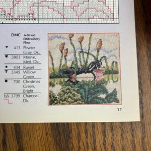May include: A cross-stitch pattern featuring a detailed image of a duck in a pond with cattails. The color palette includes shades of green, brown, pink, and blue. The pattern is on a white background with a list of DMC embroidery floss colors.