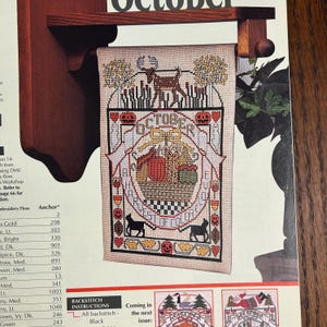 May include: A cross-stitch tapestry with the word "OCTOBER" at the top. The design features a deer, pumpkins, and the text "HARVEST OF COLORS". Two black cats are at the bottom. The tapestry is hanging on a wooden rack.