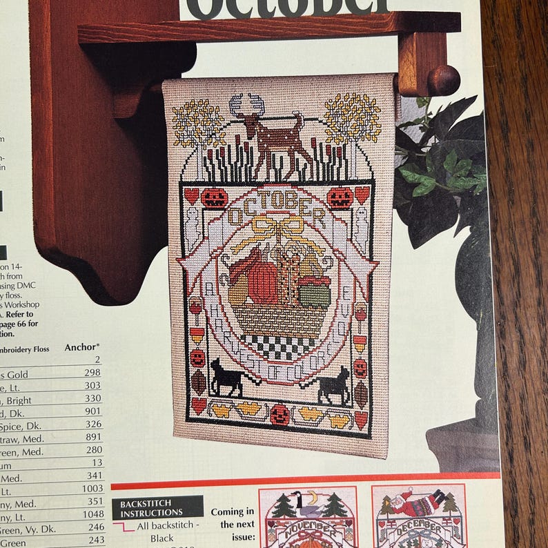 May include: A cross-stitch tapestry with the word "OCTOBER" at the top. The design features a deer, pumpkins, and the text "HARVEST OF COLORS". Two black cats are at the bottom. The tapestry is hanging on a wooden rack.