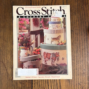 May include: A vintage issue of "Cross Stitch & Country Crafts" magazine. The cover features a stack of decorative round boxes with cross-stitched animal designs, including a cow, goat, and dog. The magazine has a floral arrangement and the text "25 Great Projects Inside!"
