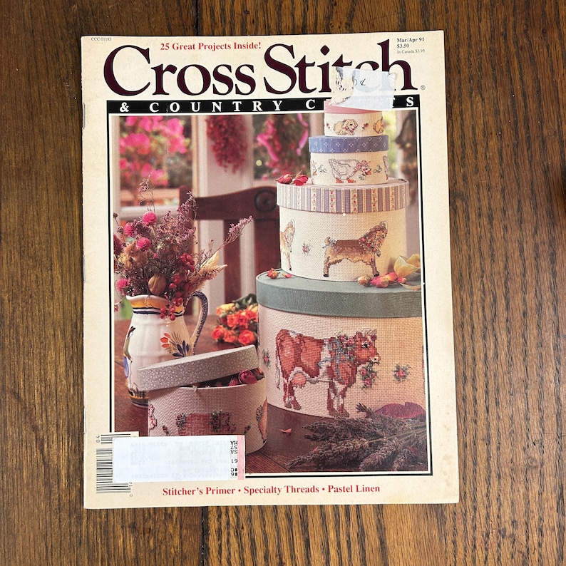May include: A vintage issue of "Cross Stitch & Country Crafts" magazine. The cover features a stack of decorative round boxes with cross-stitched animal designs, including a cow, goat, and dog. The magazine has a floral arrangement and the text "25 Great Projects Inside!"