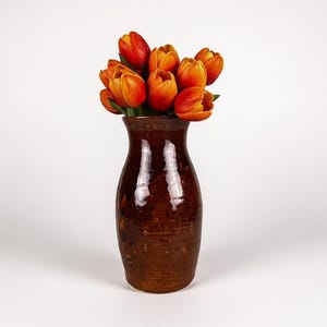 May include: A brown ceramic vase filled with orange tulips. The vase has a glossy finish and a slightly textured surface. The tulips are in full bloom, with vibrant orange petals.