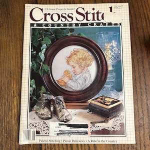 May include: A vintage issue of "Cross Stitch & Country Crafts" magazine. The cover features a framed cross-stitch of a child with a chick, a glass candle holder, and a small decorative box. The magazine's title and issue details are visible.