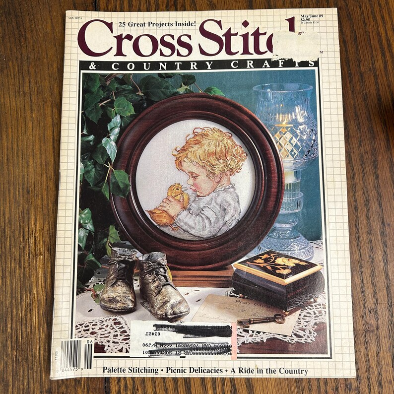 May include: A vintage issue of "Cross Stitch & Country Crafts" magazine. The cover features a framed cross-stitch of a child with a chick, a glass candle holder, and a small decorative box. The magazine's title and issue details are visible.