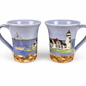 May include: Two ceramic mugs with blue handles and rims. Each mug features a coastal scene with a lighthouse and a sailboat. The base of each mug has a textured, brown, rocky design. The mugs are painted with blue, yellow, white, and brown colors.
